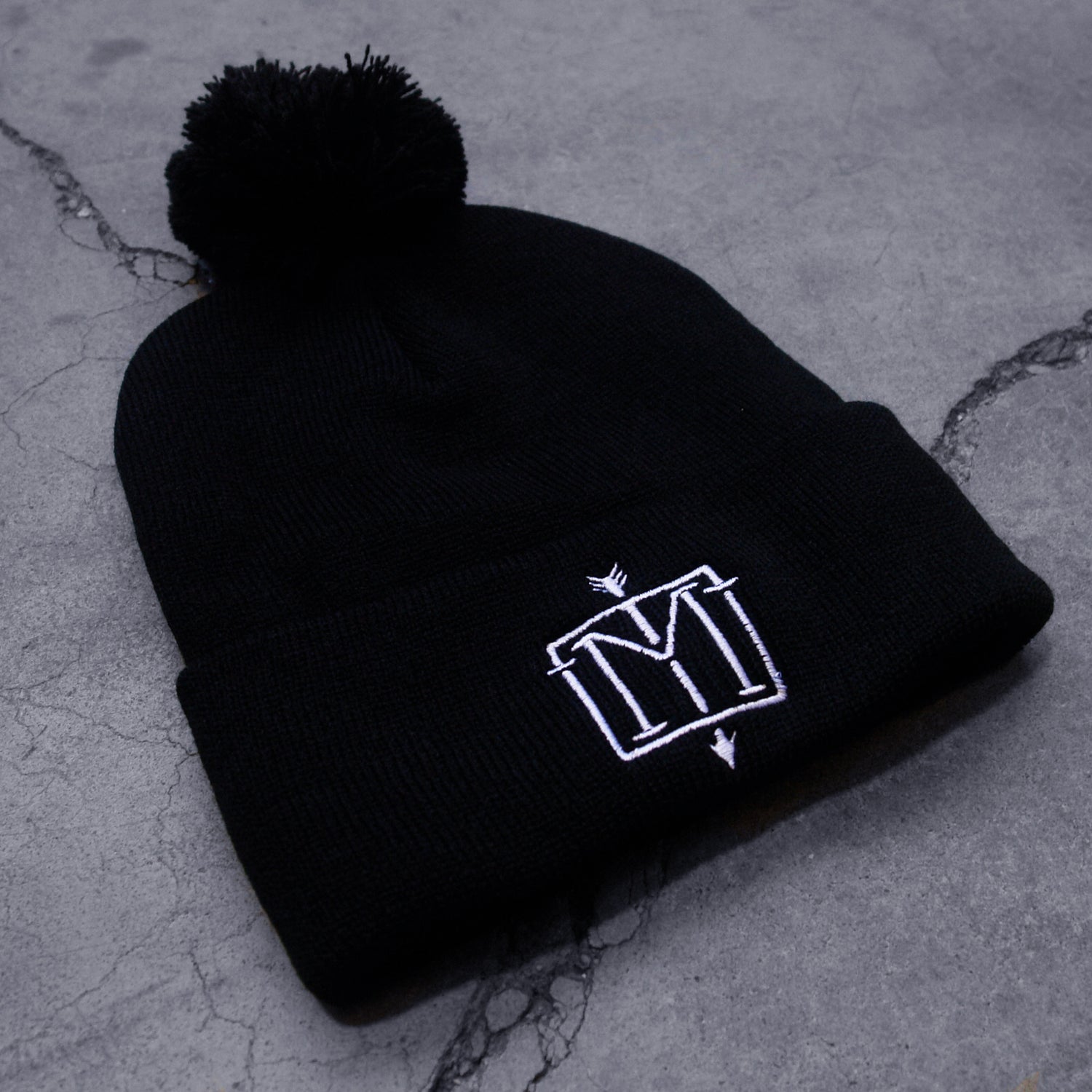 angled close up image of black winter pom beanie on cracked cement background. top of beanie has puff ball and the cuff of the beanie has in the center a white embroiderd emblem M with an arrow down the center. 