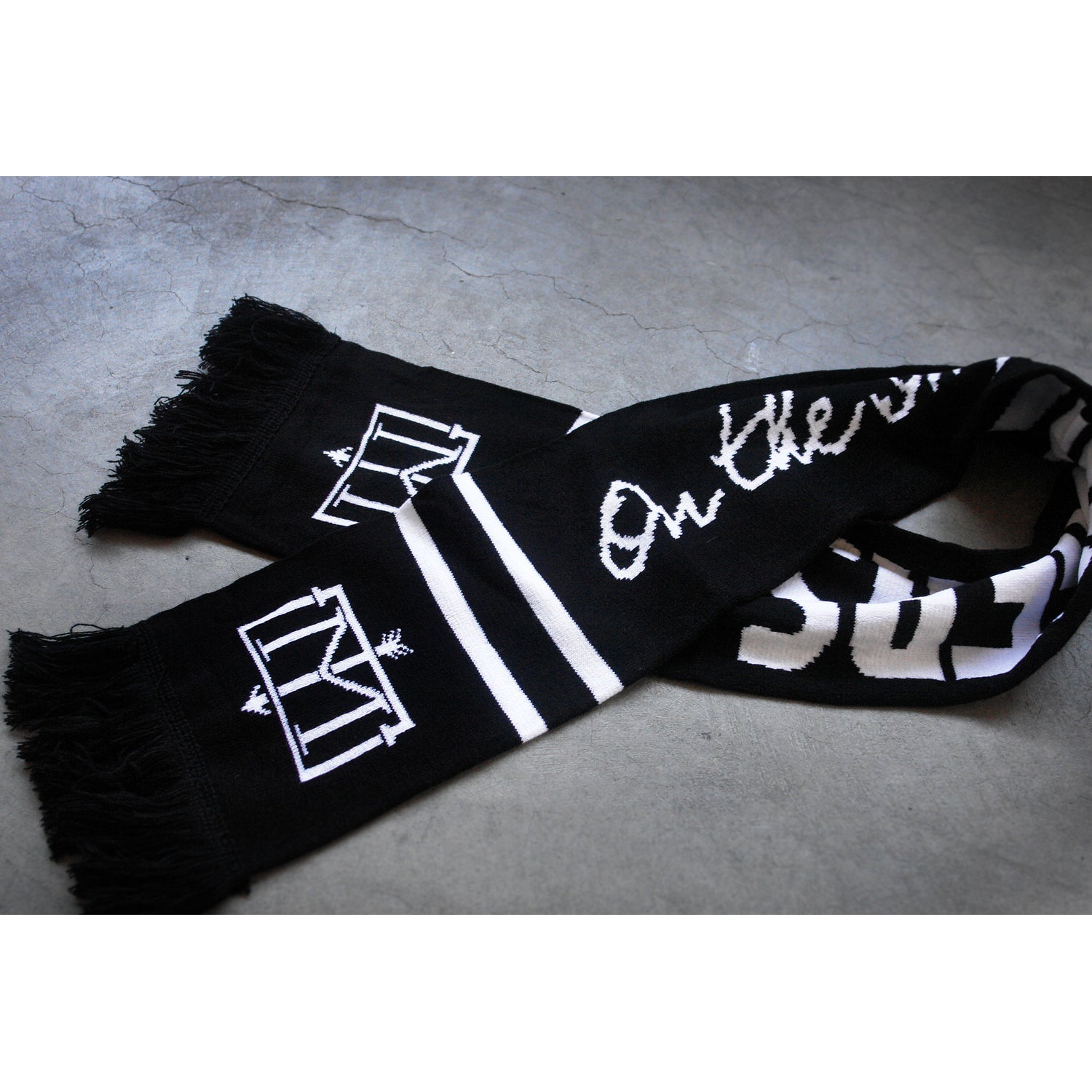 folded black and white scarf on a concrete floor. Menzingers T M emblem on the end next to the fringe