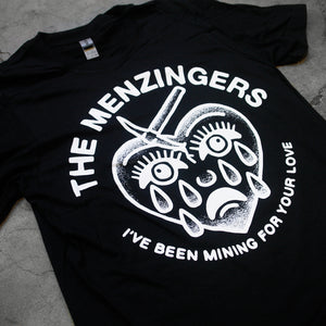 angled close up photo of black tee laid flat on cement floor. tee has full chest print in white on the front that has arched text that says the menzingers on the top and arched up text below that says i've been mining for your love. between the text is a drawing in white of a heart with a face that has a spike in the top left of the head and tears down the face.