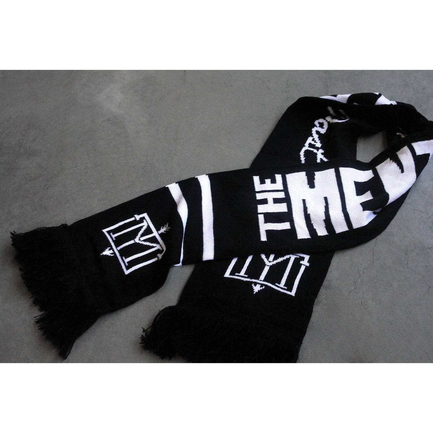 folded black and white scarf on a concrete floor. Menzingers T M emblem on the end next to the fringe