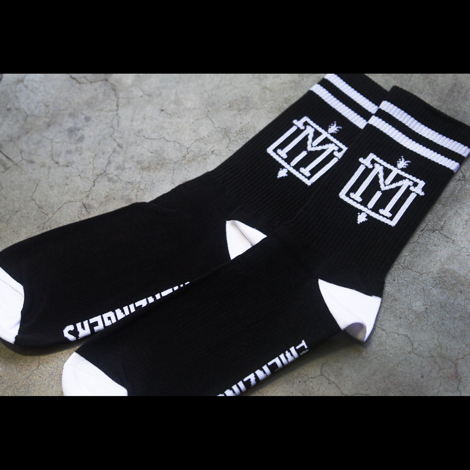 close up angled photo of two knit socks on cement background. image of two socks on white background. socks are black with two white stripes at the top and white toes and heals. there is a white emblem with the letter M and an arrow down the center on the part of the sock that is above the ankle and it says the menzingers in white on the side bottom of the socks.
