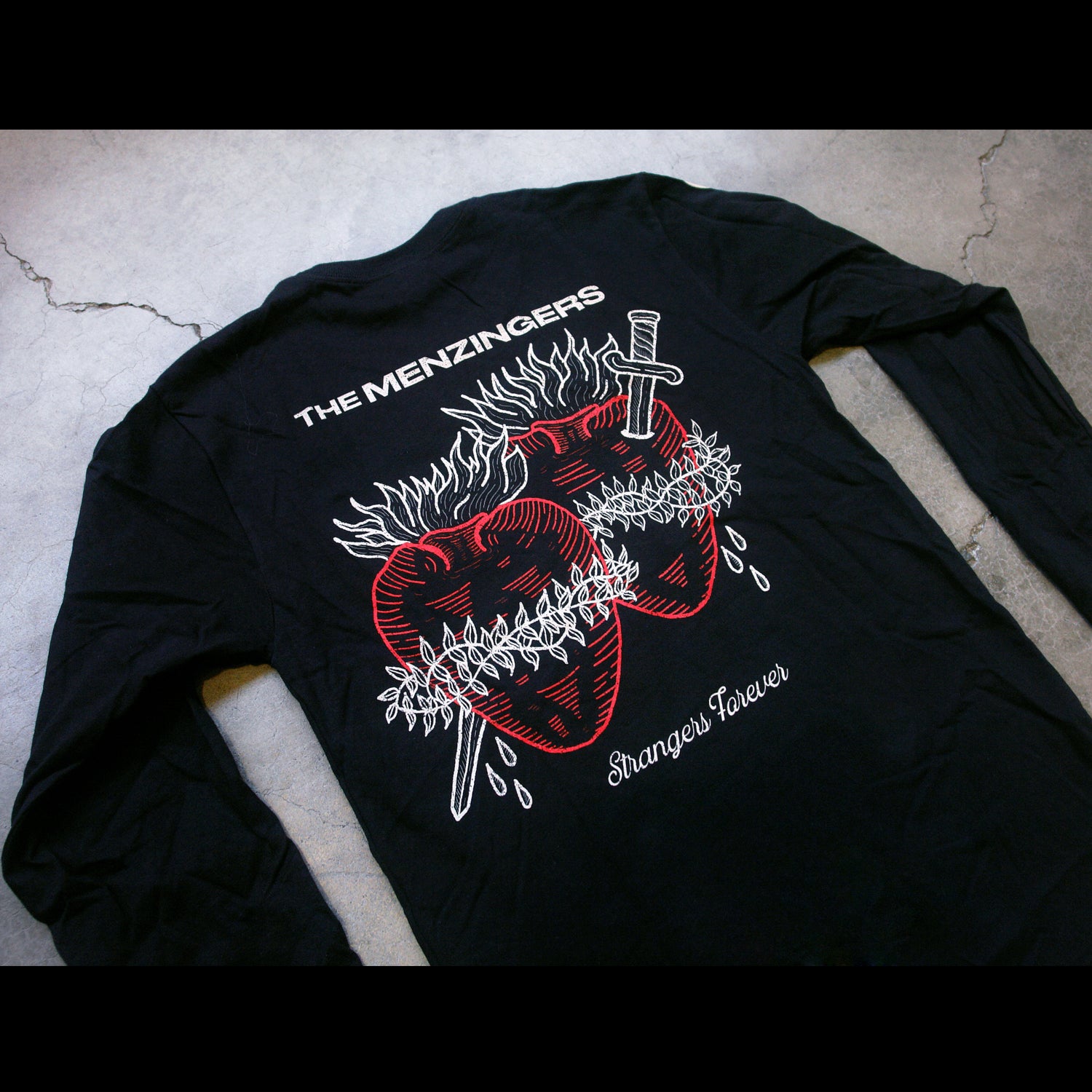 angled photo of the back of a long sleeve tee laid flat on a cracked concrete floor. full back print with cream on top that says the menzingers & two hearts in red with a sword through them. the hearts have cream fire on top barb wire wrapped around. they are bleeding in cursive below strangers forever.