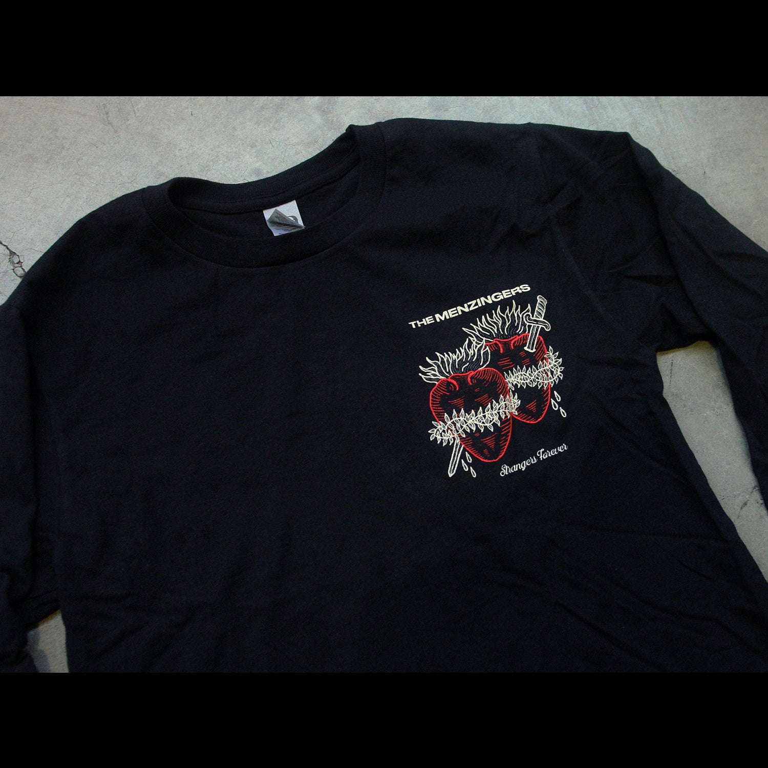 close up angled image of the front of a black long sleeve tee's chest print on the right. cream on top that says the menzingers & two hearts in red with a sword through them. the hearts have cream fire on top barb wire wrapped around. they are bleeding in cursive below strangers forever.