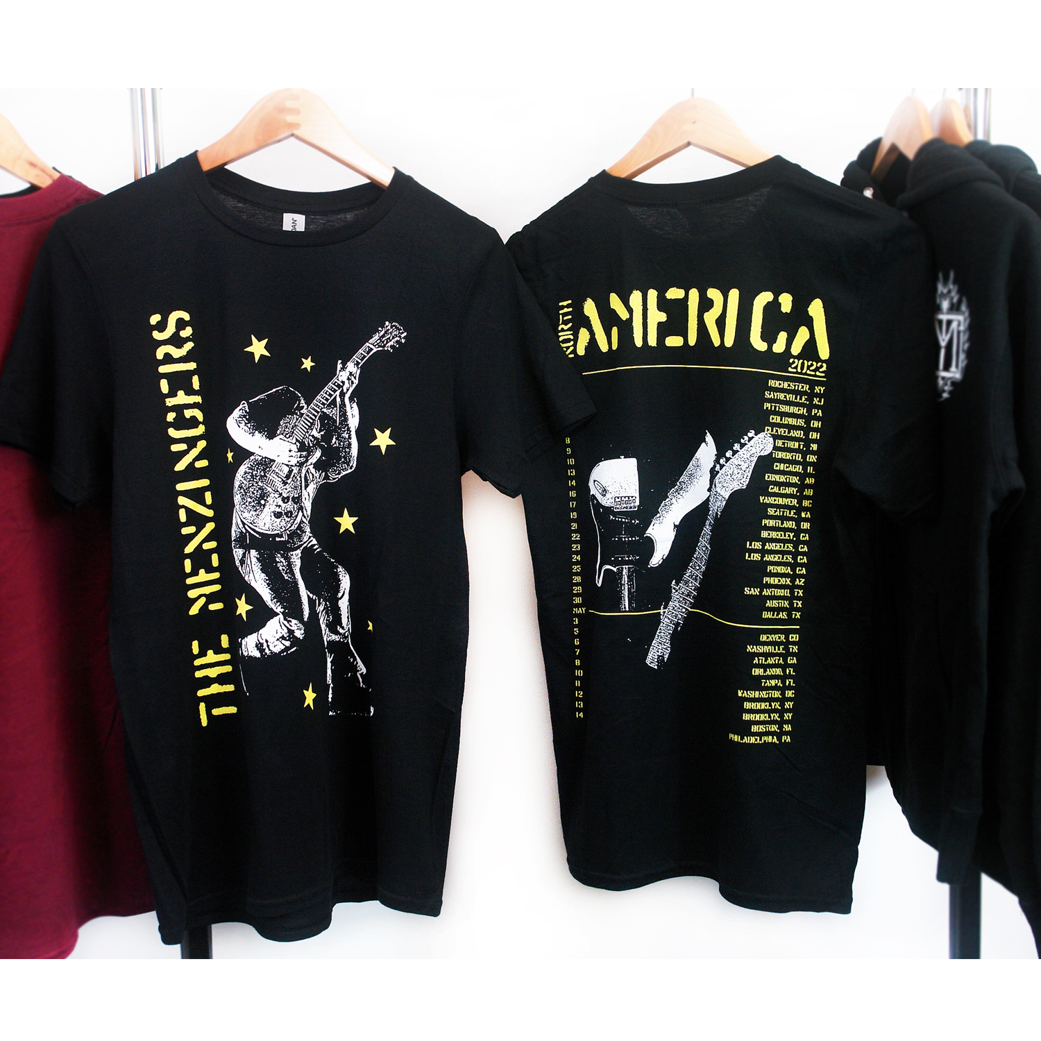 image of the front and back of a black tee shirt hanging on a white background. front of tee is on the left and has a full body print in yellow on the left side vertically says the menzingers with yellow stars all over and in white an image of a man playing a guitar. the back of the tee is on the right and has in yellow at the top 2022 america with the list of tour dates and locations below and a broken guitar in the center